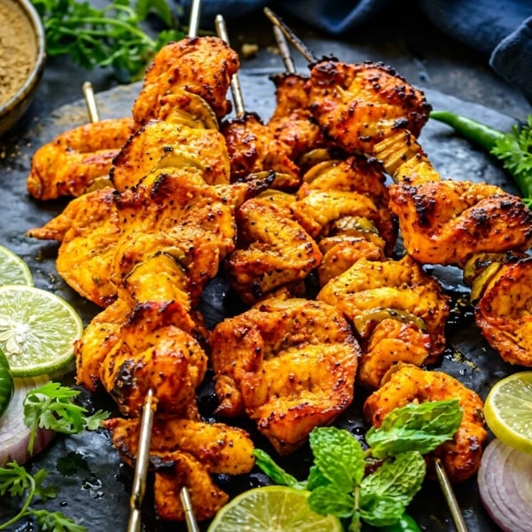 Grilled chicken tikka skewers with lime, red onion, green chili, and mint on a dark platter.